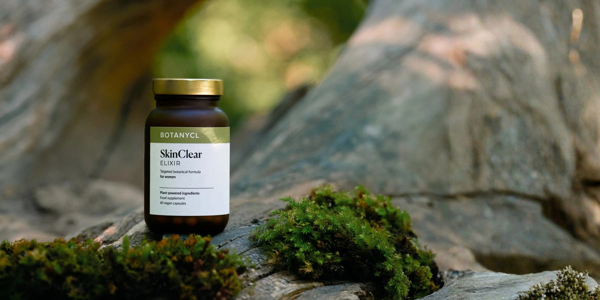 A bottle of Botanycl SkinClear Elixir skin supplement standing on mossy rocks in an outdoor forest setting.